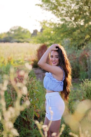 Smiling young woman with long wavy hair poses in a blue ruffled top and white skirt in a sunlit field at golden hour.