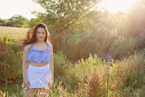 Young woman in a blue striped crop top and white skirt stands among wildflowers and tall grass in warm evening sunlight.