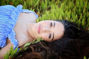 Close-up of a woman with dark hair lying in green succulent plants, wearing a blue striped top and gazing at the camera.