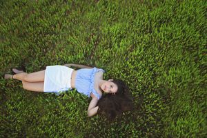 Overhead view of a young woman in a blue top and white skirt lying on lush green grass with her long hair spread out.