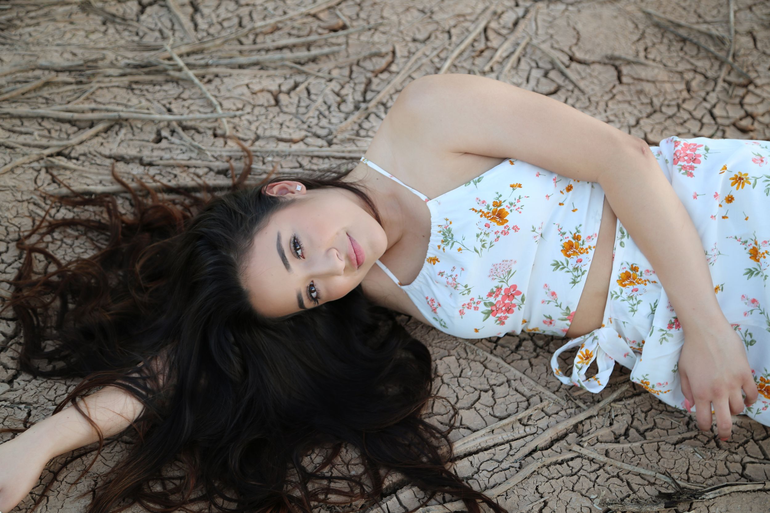 Young woman with long dark hair lies on cracked dry earth wearing a white floral sundress and smiling softly at the camera.