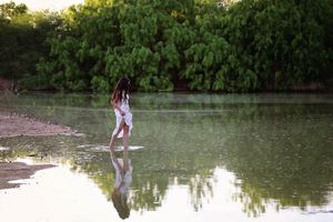 Woman in a white dress walks barefoot through shallow lake water, her reflection mirrored on the calm surface beside green trees.