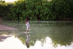 Woman in a light dress wades through calm shallow water near the shore, with her reflection and green trees in the background.