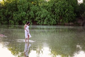 Young woman in a white dress stands in a still lake creating gentle ripples, with lush green trees and a soft reflection behind her.