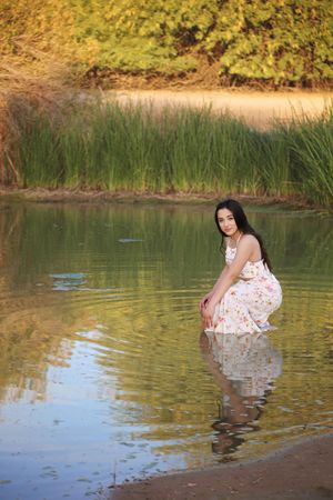 Woman in a floral sundress crouches at the edge of a reflective pond with tall reeds and golden foliage in the background.
