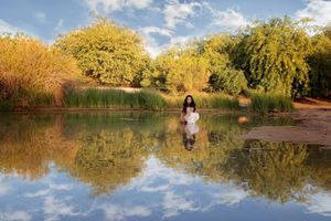 Woman in a white dress sits at a pond's edge, her reflection mirrored in the water beneath autumn trees and a blue sky.