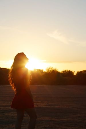 Silhouette of a woman with long hair in a red dress standing in an open field as the golden sun sets behind her.