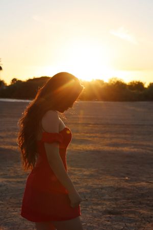 Woman in a red dress looks down while golden sunset light creates a warm glow and lens flare across an open field.