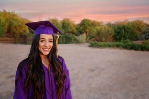 Smiling graduate with long dark hair wears a purple cap and gown outdoors with a colorful sunset sky behind her.