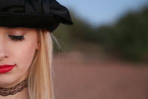 Close-up portrait of a woman with blonde hair, red lipstick, a lace choker, and a black bow hat looking downward outdoors.