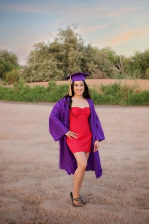 Graduate in a purple cap and open gown over a red dress poses with a smile on dry ground near green shrubs at dusk.