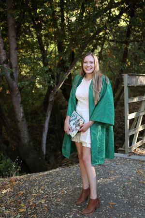 Smiling graduate in a green gown over a white dress and brown boots holds a decorated cap near trees and a wooden fence.