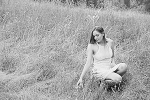 Black and white photo of a woman in a light dress sitting in tall grass on a hillside, gently touching the blades.