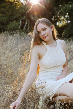 Young woman with long hair in a cream lace dress sits in golden dry grass as sunlight streams through the trees behind her.