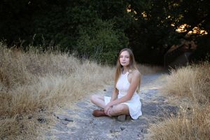 Young woman in a white sundress sitting cross-legged on a dirt path surrounded by dry golden grass and dark trees.