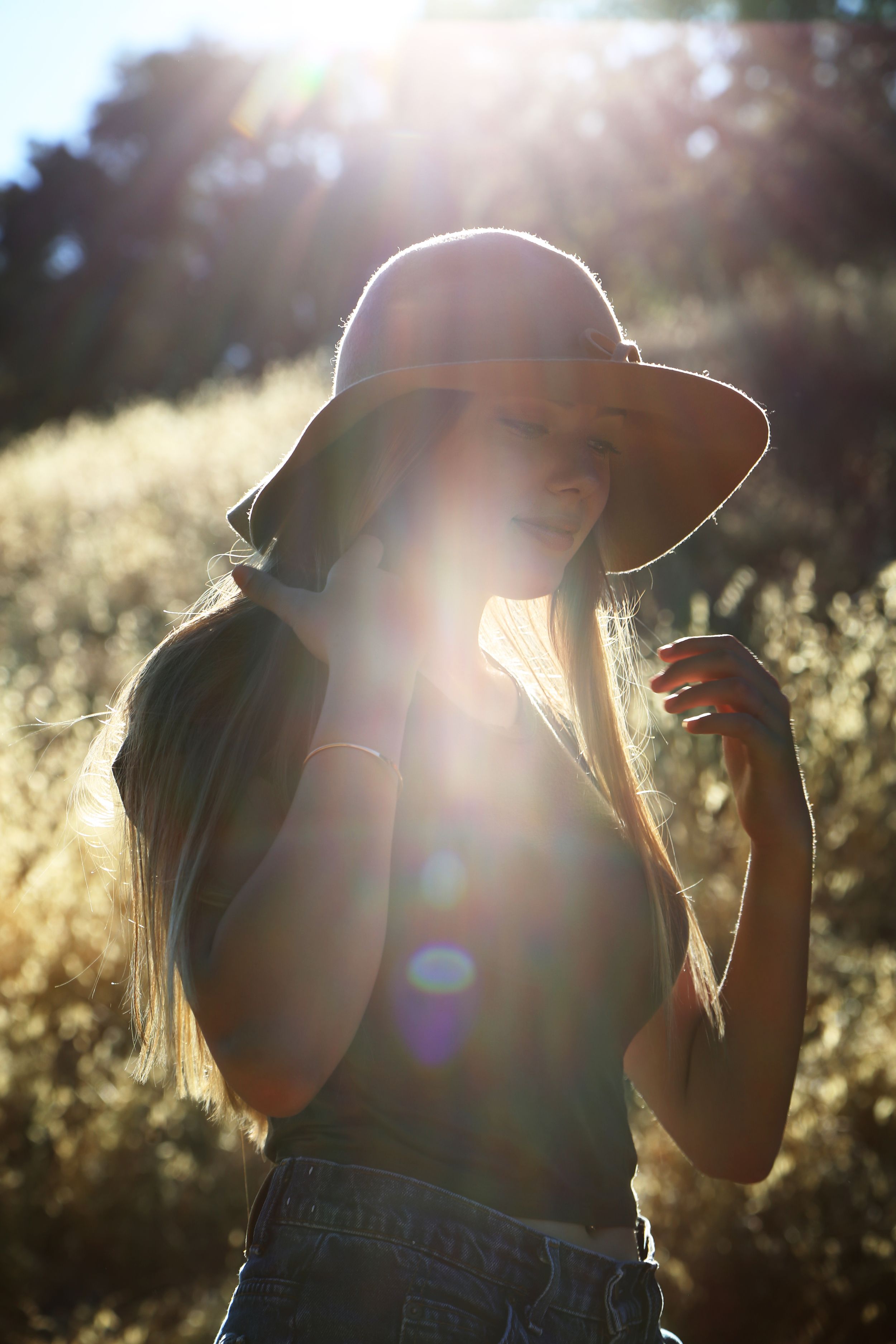 Backlit woman in a wide-brim hat and denim shorts standing in glowing golden sunlight with dramatic lens flare.