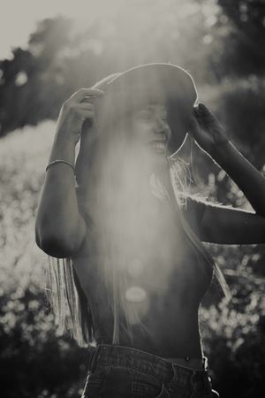 Monochrome image of a laughing woman holding her wide-brim hat, long hair flowing, with bright sun flare through the haze.