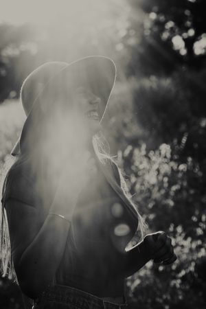 Black and white backlit photo of a smiling woman in a brimmed hat with sun flare and blurred foliage behind her.