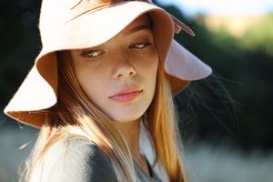 Close-up portrait of a young woman with long hair wearing a blush wide-brim floppy hat in soft natural outdoor light.