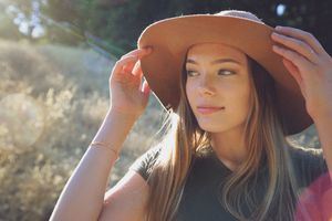 A young woman in a wide-brimmed hat adjusts it with both hands while gazing softly aside, lit by warm golden sunlight outdoors.