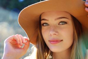 Young woman with light eyes in a tan wide-brim hat gently holds the brim while smiling softly in warm golden sunlight.