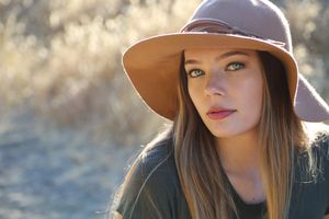 Portrait of a young woman with green eyes and long hair wearing a tan wide-brim hat against a softly blurred golden field.