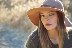 Close-up portrait of a young woman with long hair and green eyes wearing a wide-brimmed tan hat in warm, golden sunlight.