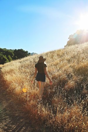 A woman in a hat and shorts walks uphill through tall golden grass under a bright blue sky with a sun flare.