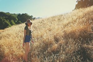 A woman in a brown hat and denim shorts stands on a golden grassy hillside with green trees and a warm, hazy sky behind her.
