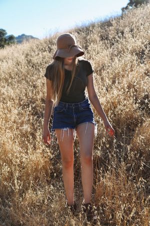 Young woman in a brown floppy hat, black tee and denim cutoff shorts walks through golden dry grass on a sunlit hillside.