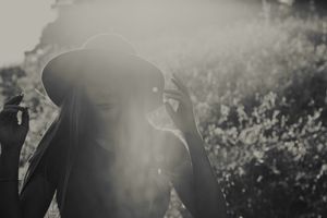 Black and white photo of a woman adjusting her dark brimmed hat in hazy sunlight with a softly blurred leafy background.