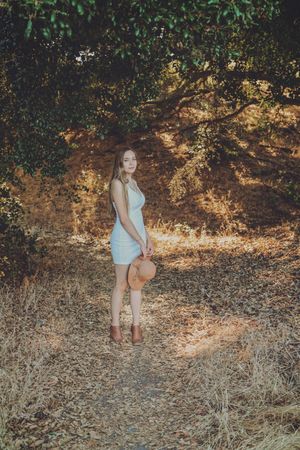 Young woman in a light blue dress and brown boots holds a straw hat while standing on a dry leaf-covered path beneath oak trees.