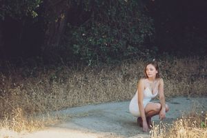 A young woman in a white dress crouches on a dirt path beside dry grass, with dark green trees in the background.