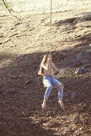 A young woman in ripped jeans swings barefoot on a rope over a sunlit ground covered in dry leaves and rocks.