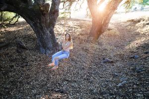 Woman in jeans sitting on a rope swing hanging from an oak tree, with sunlight streaming through the branches onto the leaf-covered ground.