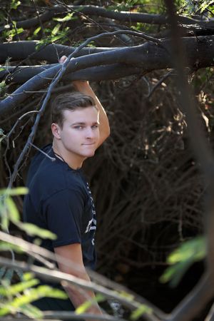 Young man with short brown hair in a black t-shirt stands among fallen tree branches, looking at the camera in dappled sunlight.