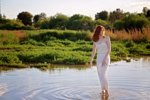 Red-haired woman in a flowing white dress wades through shallow water with green grass and trees in the background at sunset.
