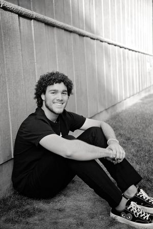 Black and white portrait of a smiling young man with curly hair sitting on grass beside a wooden fence, wearing dark clothes and sneakers.