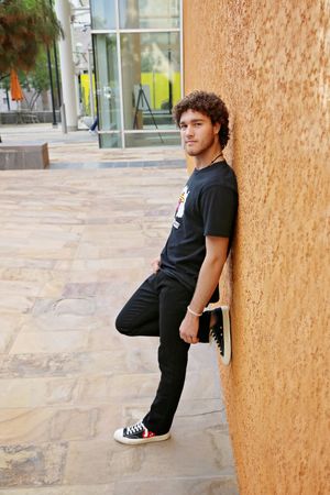 Young man with curly hair in a black t-shirt and sneakers leans casually against an orange stucco wall on a stone-paved plaza.