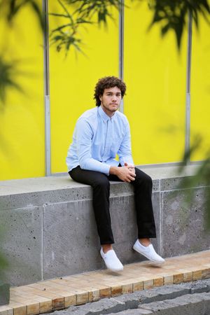 Young man with curly hair in a light blue shirt and white sneakers sits on a concrete ledge in front of a vivid yellow wall.