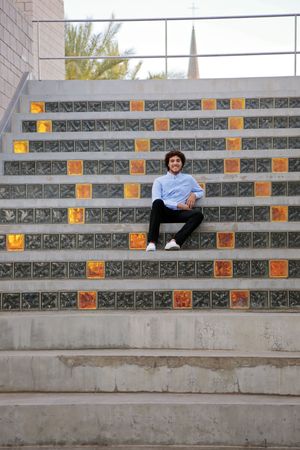 Young man in a light blue shirt sits and smiles on wide concrete steps decorated with colorful tiles, with a church steeple behind.