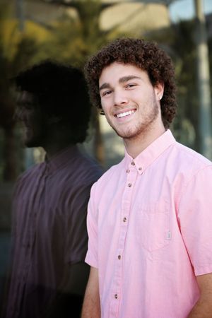Smiling young man with curly hair and light stubble wears a pink button-up shirt, with a darkened reflection visible in glass beside him.