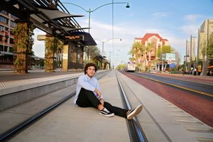 Young man in a light shirt and black pants sits on light rail tracks in a city street lined with palm trees and buildings.