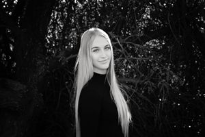 Black and white portrait of a young woman with long straight blonde hair, wearing a dark top, smiling softly against dark foliage.