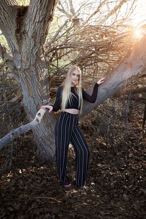 Young woman with long blonde hair in a black crop top and striped pants leaning against a large tree at sunset.