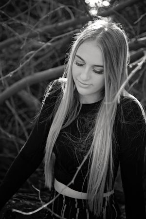 Black and white portrait of a young woman with long straight blonde hair looking down, standing among bare tree branches.