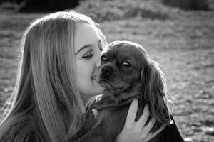 Black and white photo of a girl with long hair kissing a cavalier King Charles spaniel on the cheek outdoors.