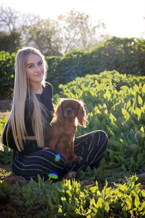 Smiling young woman with long blonde hair sitting in a green garden holding a brown cavalier King Charles spaniel.