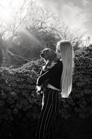 Black and white photo of a woman with long hair hugging her dog beside ivy-covered bushes with sunlight streaming through.