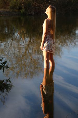 Young woman in a floral dress standing in shallow lake water at sunset, her reflection mirrored on the calm surface.